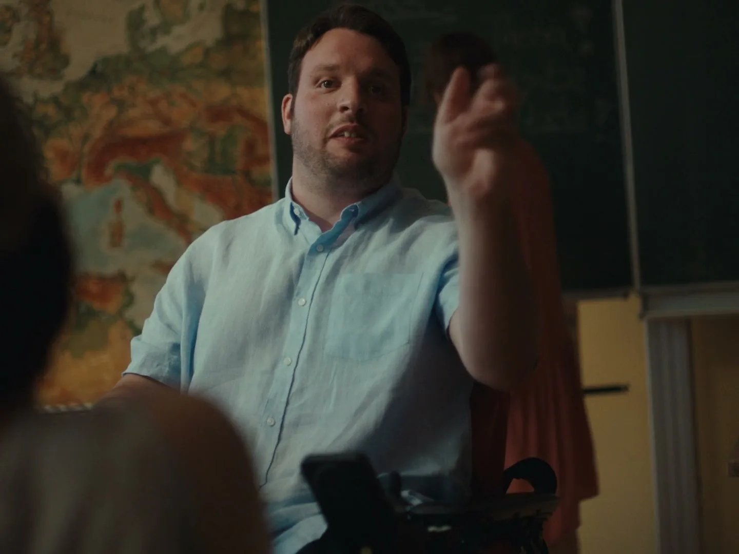 A man in a light blue shirt, sitting in a wheelchair, talks and gestures in a classroom with a map on the wall.