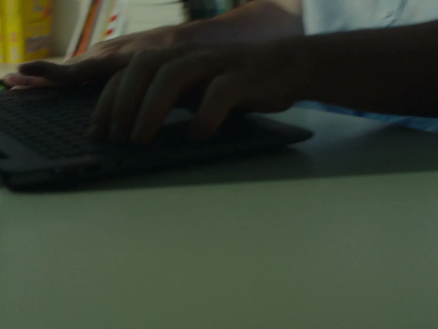 A young man in a light blue shirt types on a laptop, with bookshelves visible in the background, in an interior setting.