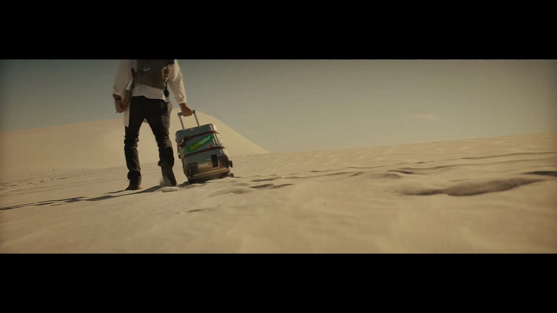 A person in a white shirt and dark pants walks through a vast desert landscape, pulling a suitcase with a Subway logo on it, leaving footprints in the sand.