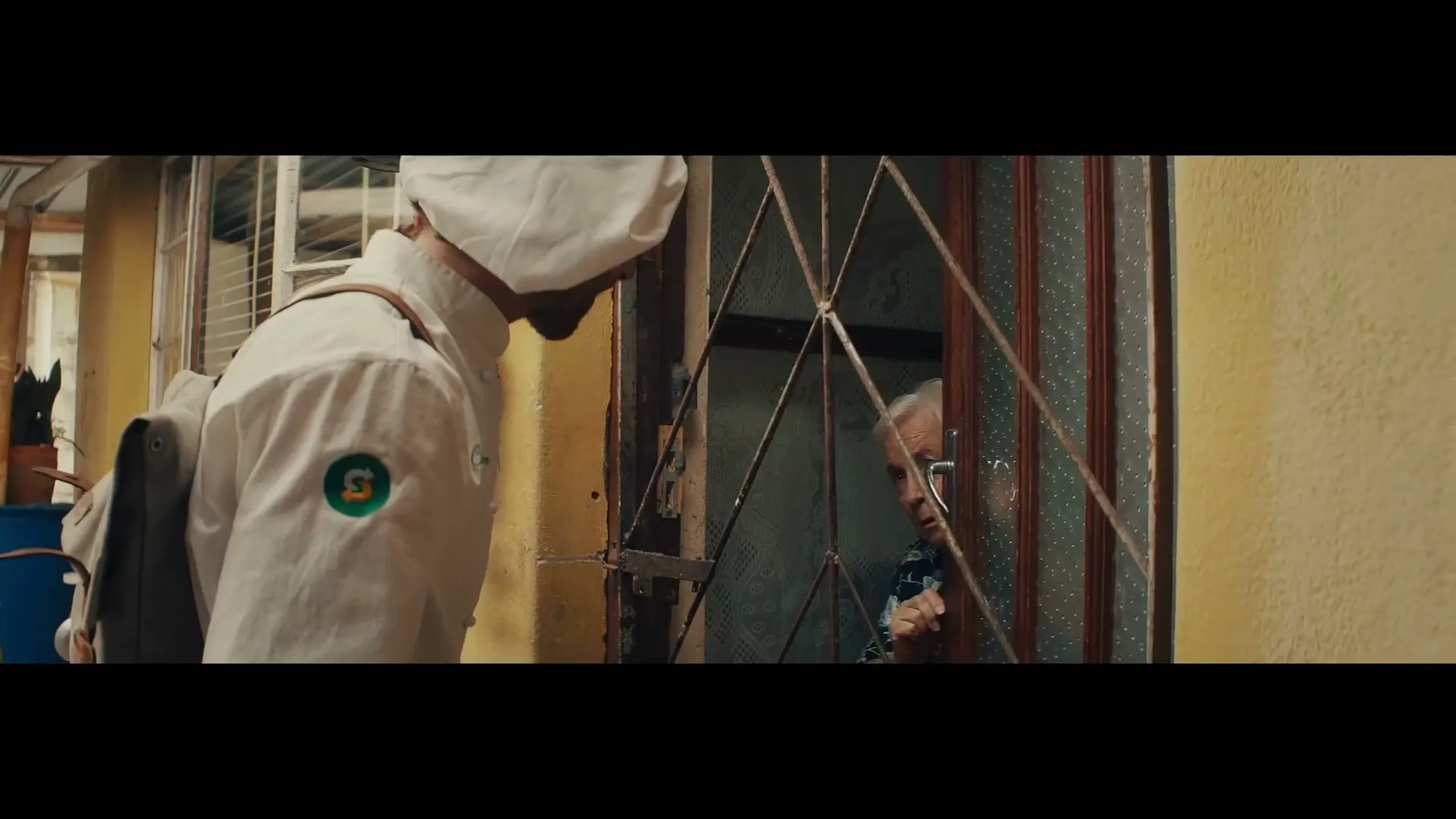 A male chef in a white uniform with a Subway logo on his sleeve looks down at an older woman through a barred window in a rustic setting.