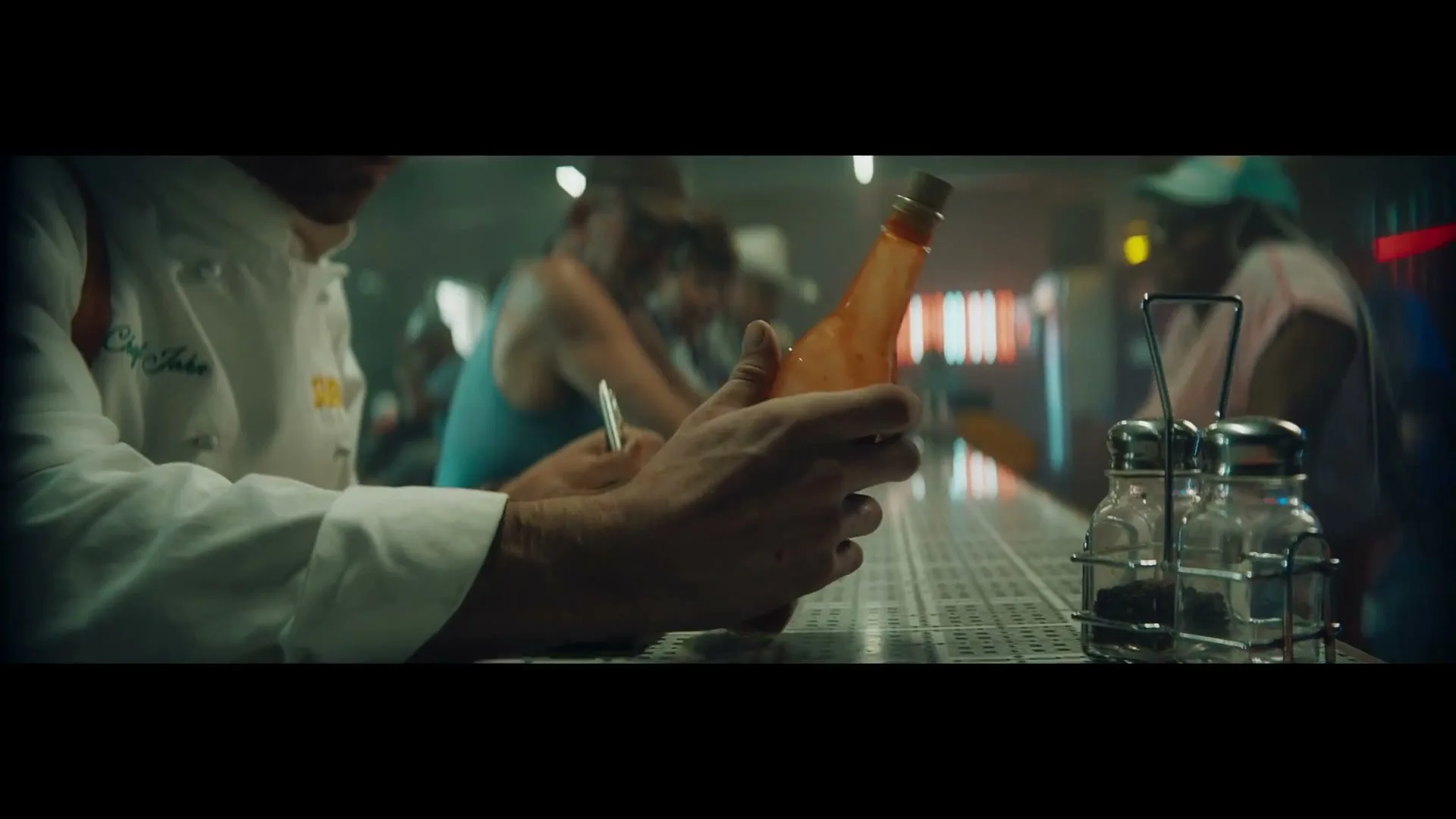 A male chef in a white uniform and hat looks at a hot sauce bottle while sitting at a diner counter.