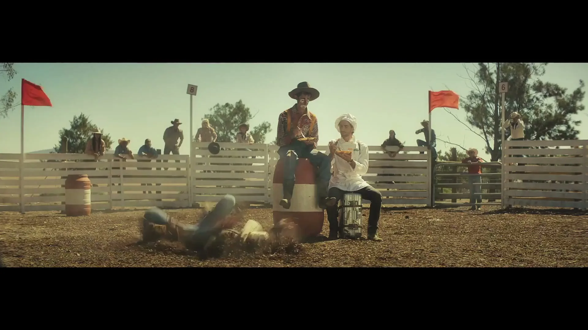 A cowboy falls off a mechanical bull while a man in a cowboy hat and a chef sit calmly eating sandwiches at a rodeo.