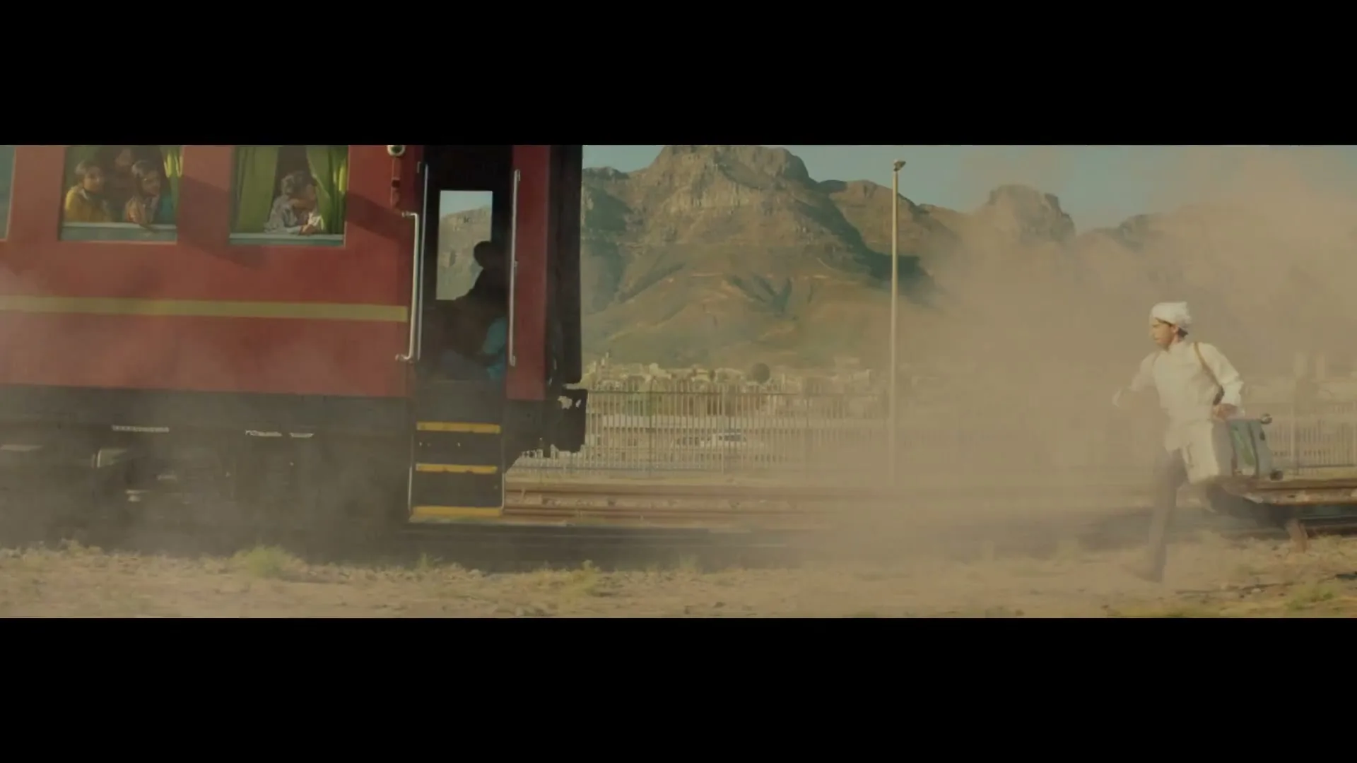 A man in a white uniform runs alongside a red train, carrying a Subway sandwich, as children look out the train windows in a dusty, mountainous landscape.