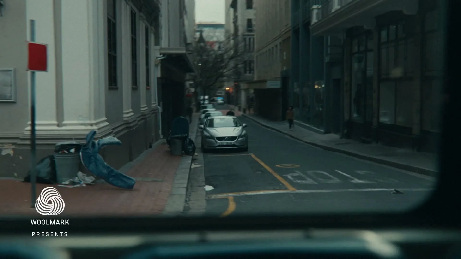 A pair of blue jeans with legs appears to run across a city street, passing a parked car and trash cans on the sidewalk.