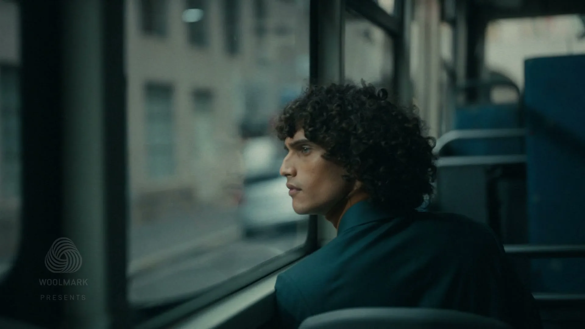 A young man with curly hair, wearing a dark teal jacket, looks out the window of a moving bus, lost in thought.