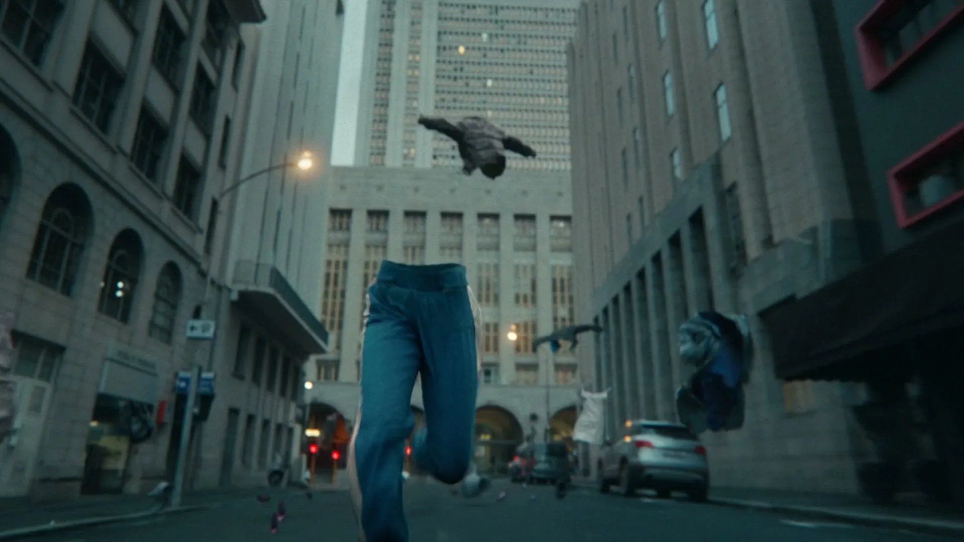 A pair of blue jeans runs down a city street, surrounded by other floating, discarded clothes, with tall buildings in the background.