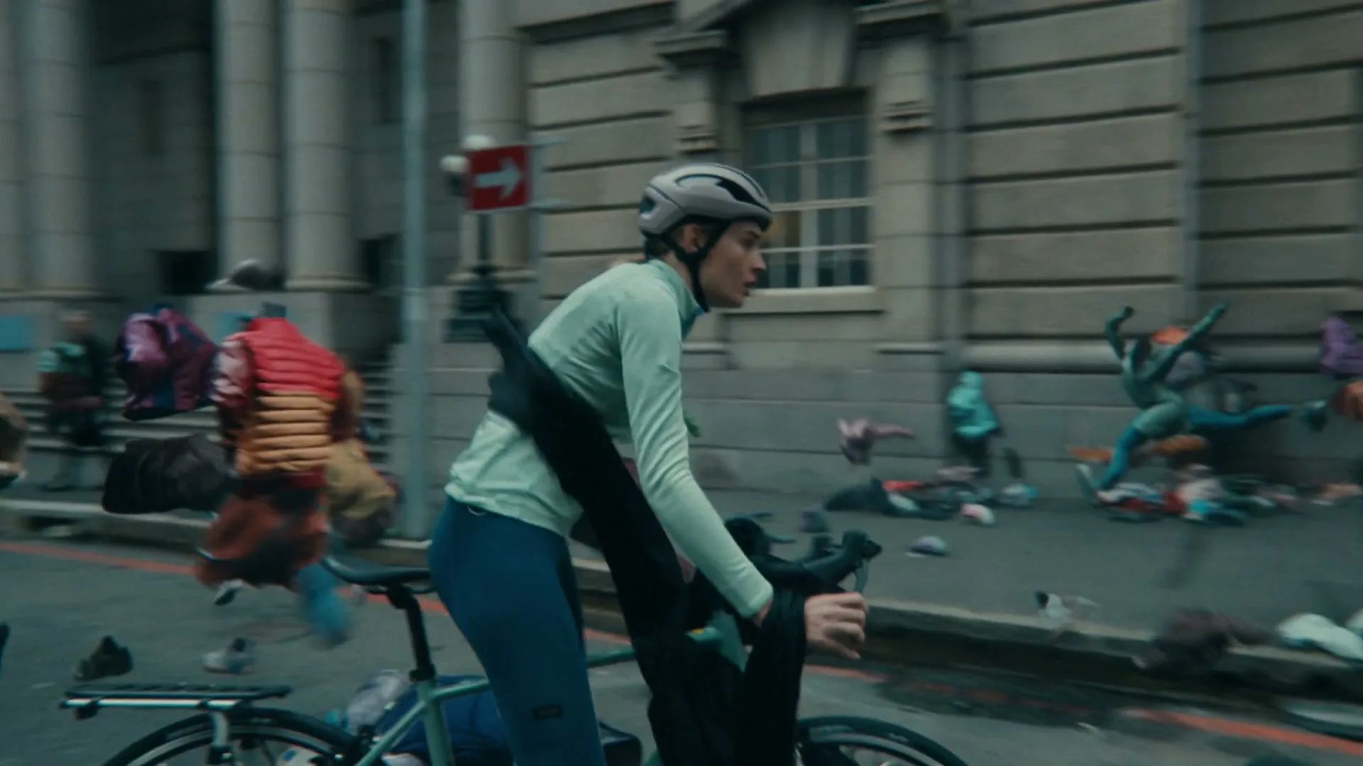 A young woman on a bicycle looks back in fear as she rides through a city street filled with discarded, animated clothing items that appear to be chasing her.