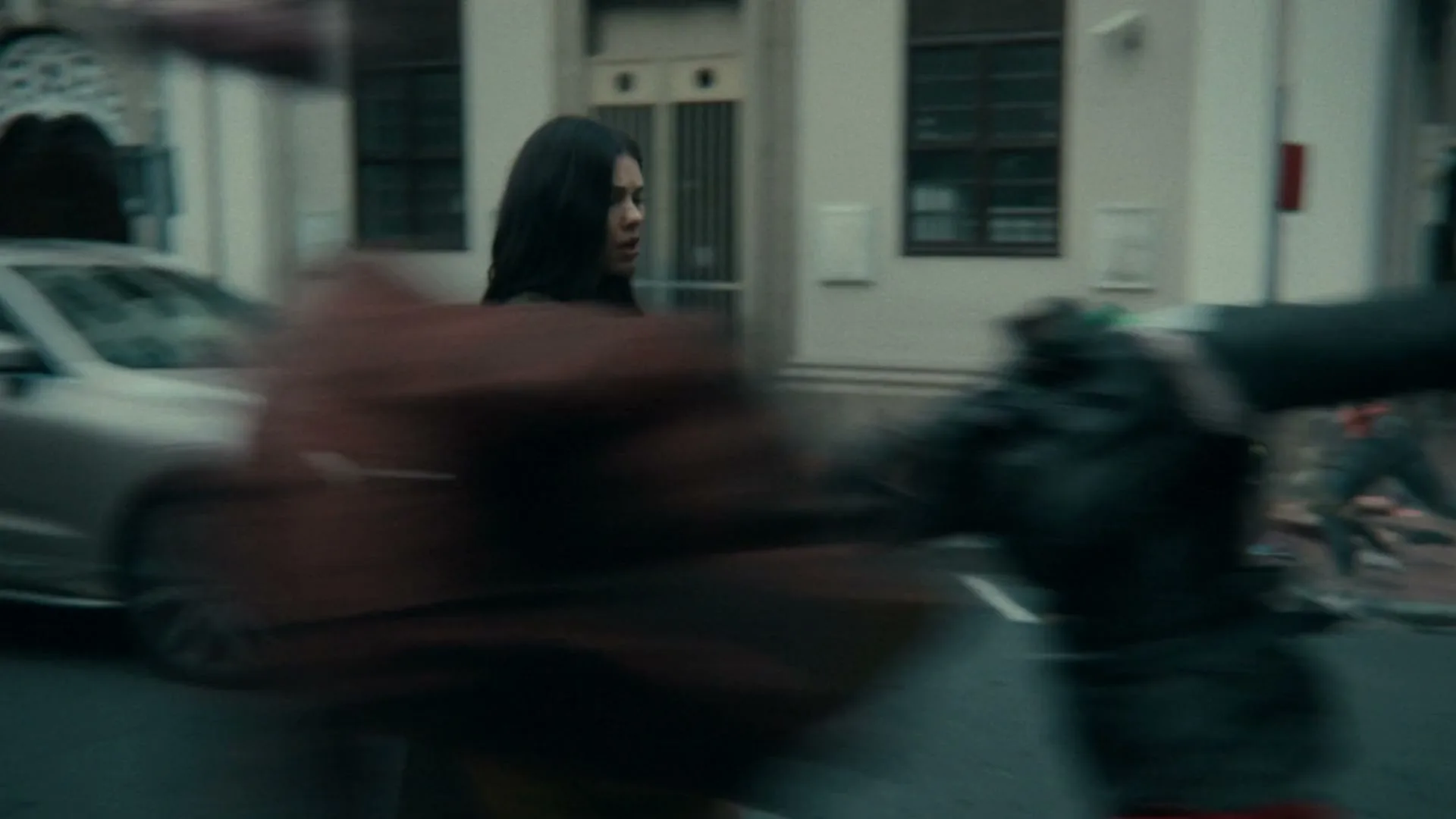 A young woman in a long coat walks through a city street as blurred, colorful clothing items fly past her, creating a sense of chaos.