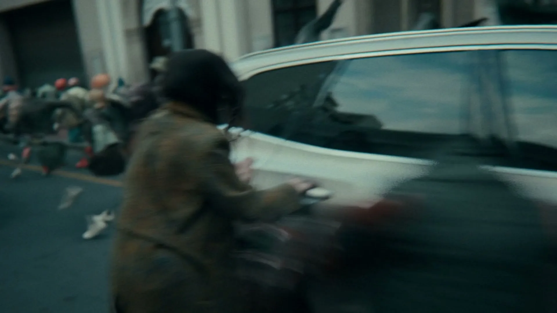 A woman in a brown coat runs towards a silver car, attempting to open the door, while other people run past her on a city street.