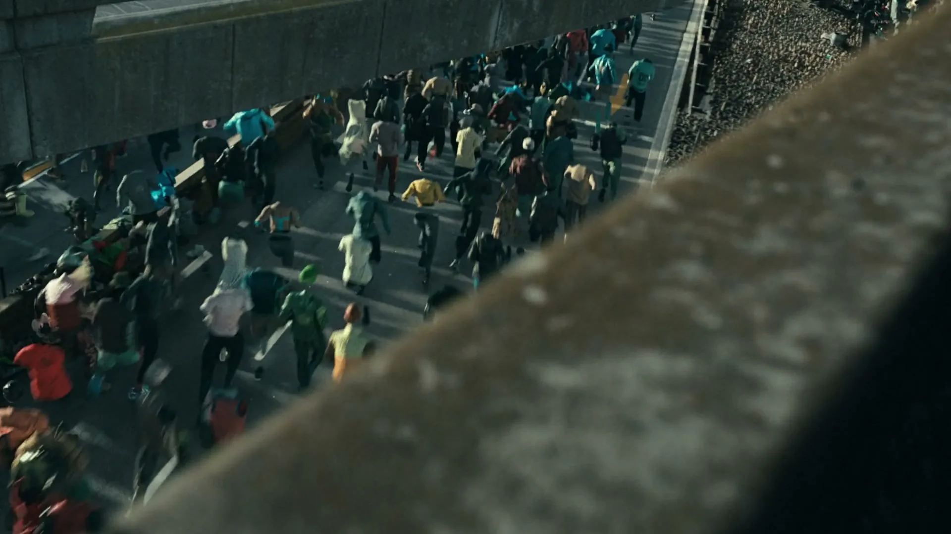 A high-angle shot shows a large crowd of people, some wearing colorful clothes, running along a concrete overpass next to a railway.
