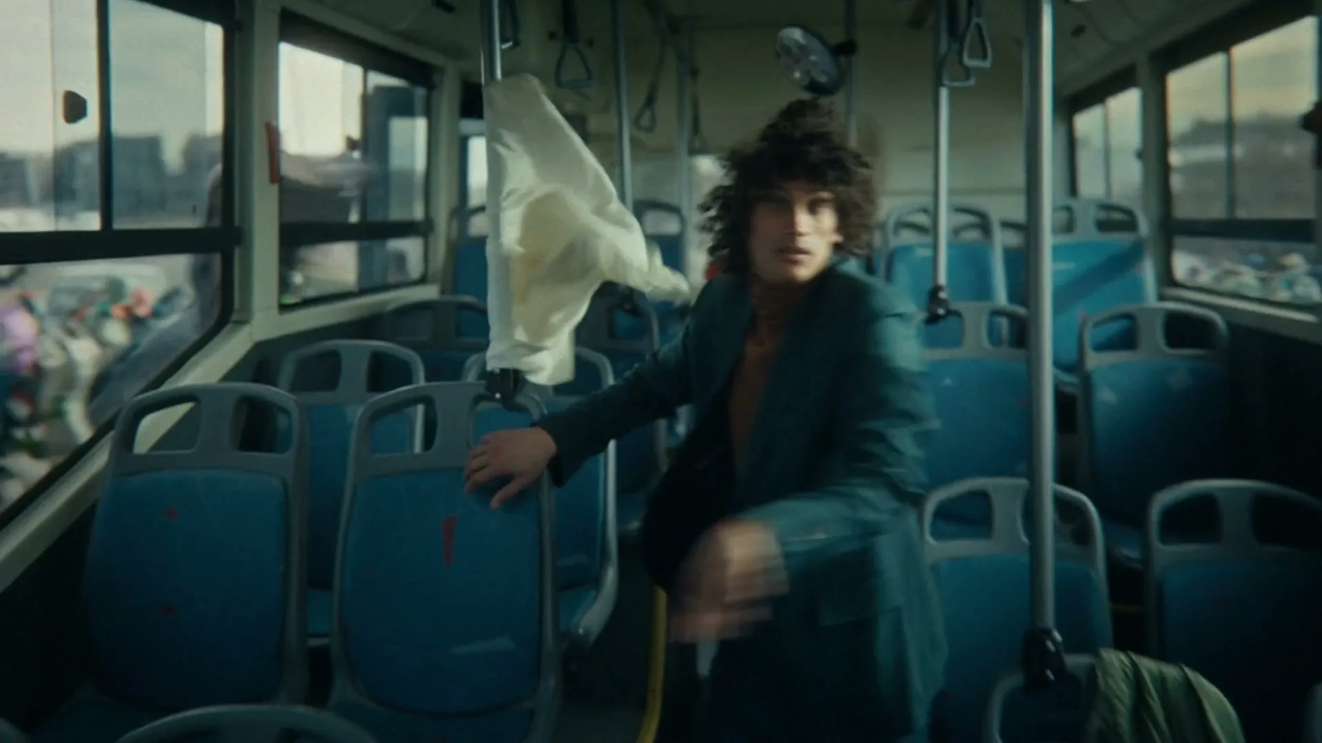 A young man with curly hair in a green suit looks surprised as he stumbles on a moving bus, with clothes flying around him.