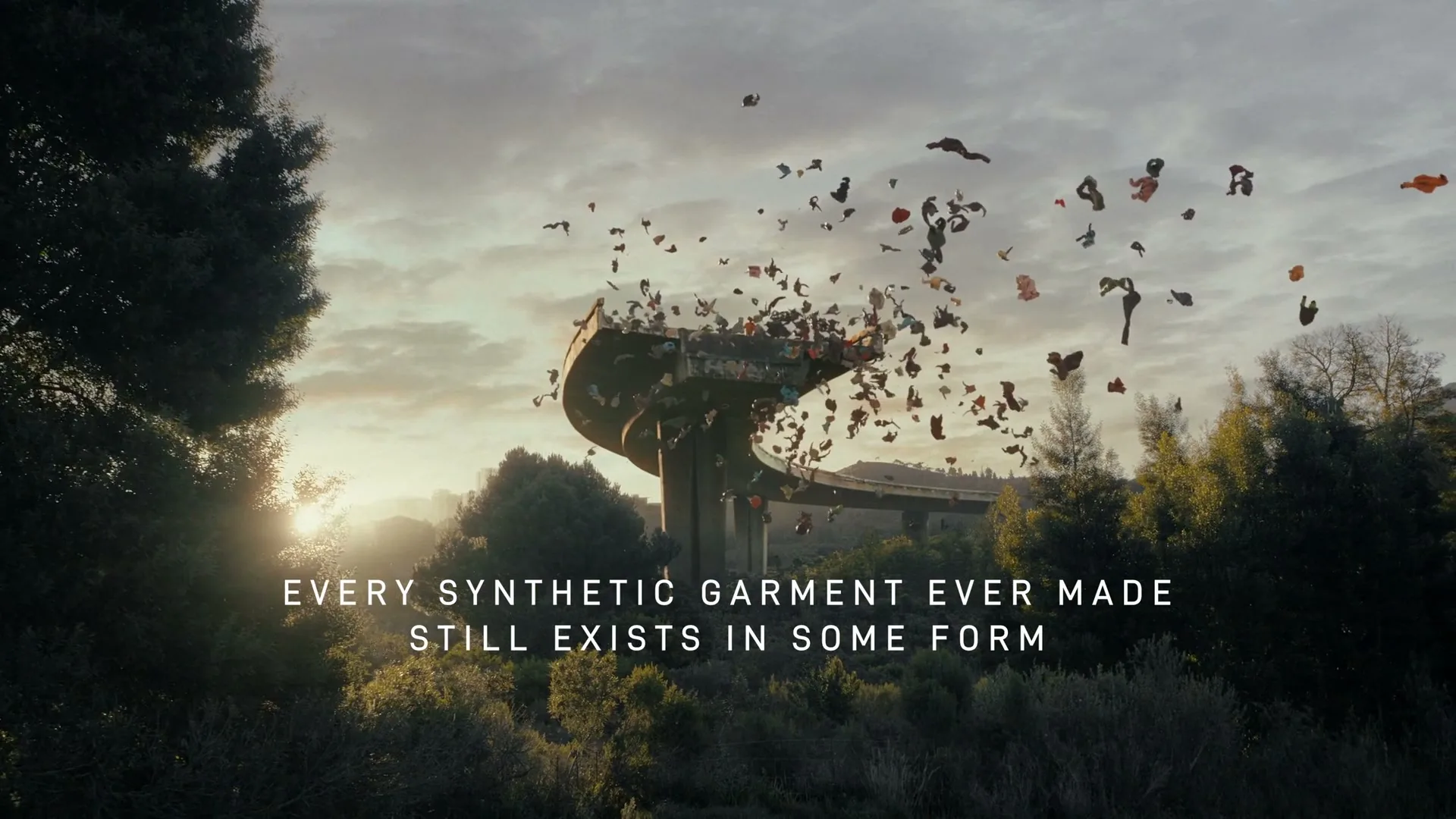 A wide shot shows a crumbling concrete overpass with thousands of pieces of clothing flying out of it, over a lush green landscape at sunset.