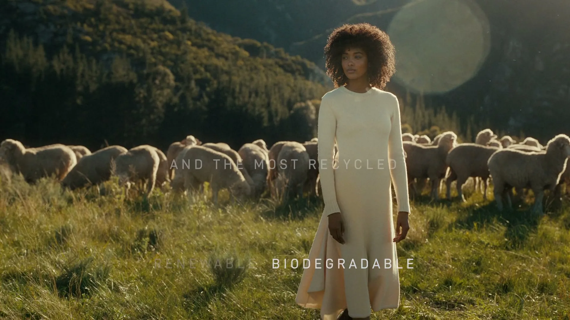 A young woman with curly hair, wearing a long cream dress, stands in a grassy field with a flock of sheep behind her, mountains in the background.