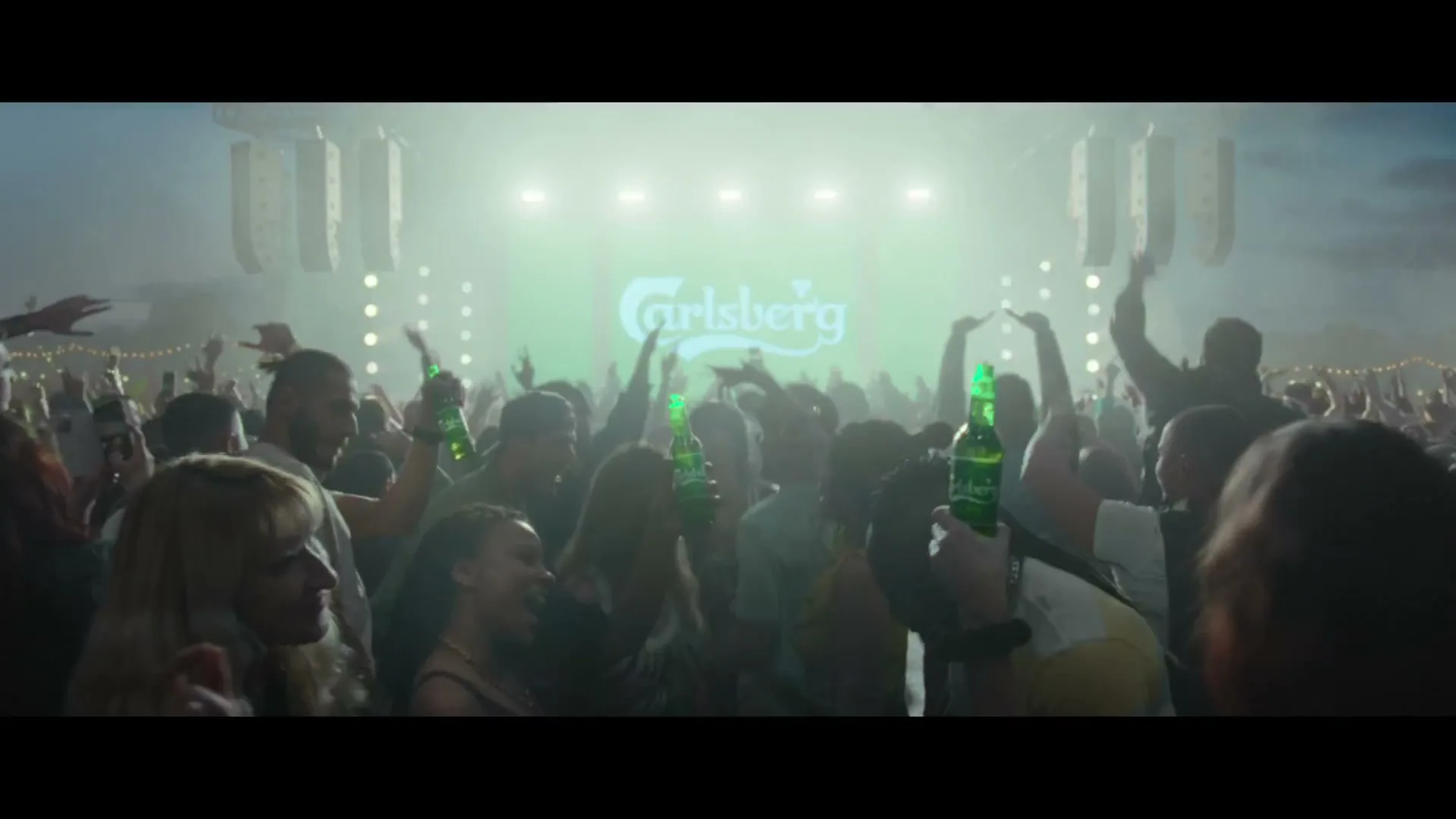 A large crowd of people at a music festival, many holding green bottles, with the Carlsberg logo displayed prominently on a large screen in the background.