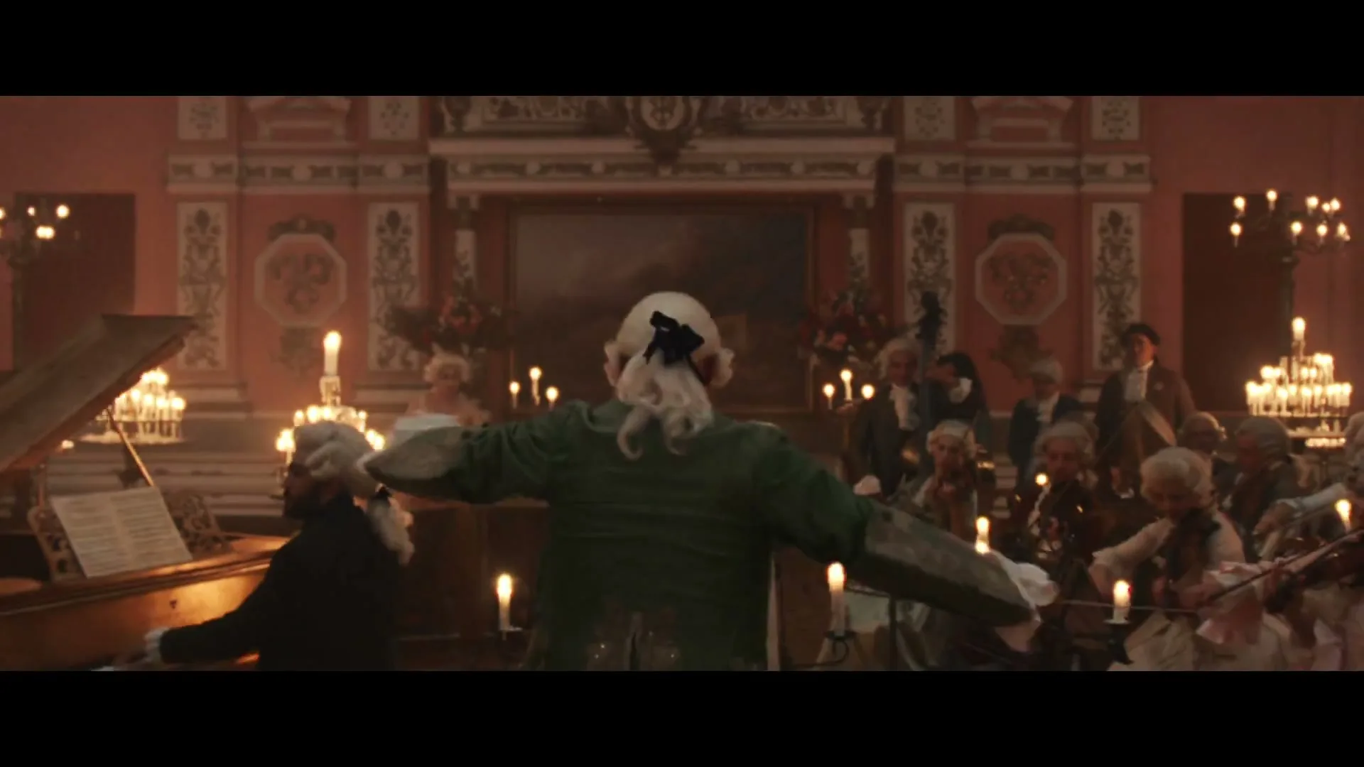 A classical orchestra with a conductor in a green coat and white wig performs in a candlelit hall, with people in period attire dancing and listening.
