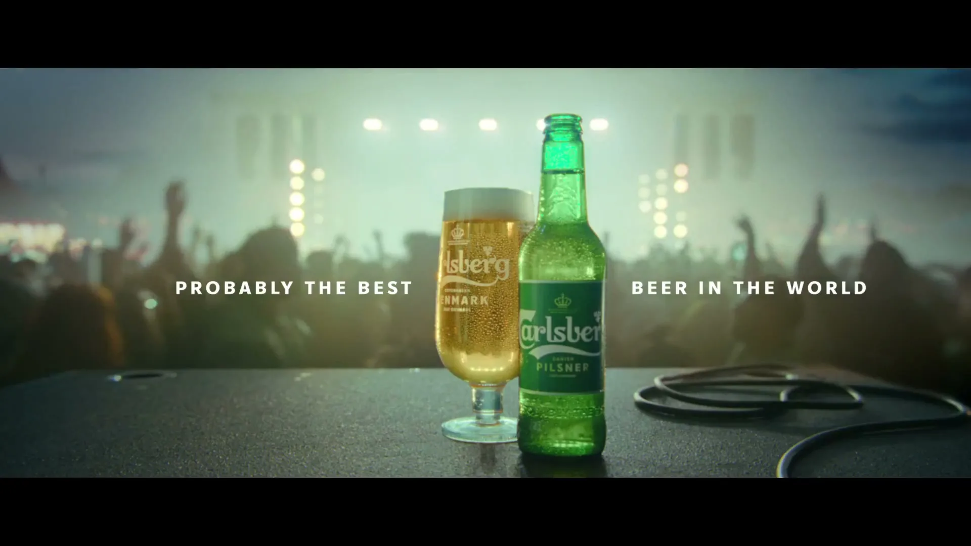 A Carlsberg beer bottle and a glass of beer are on a table in front of a blurred crowd at a concert.