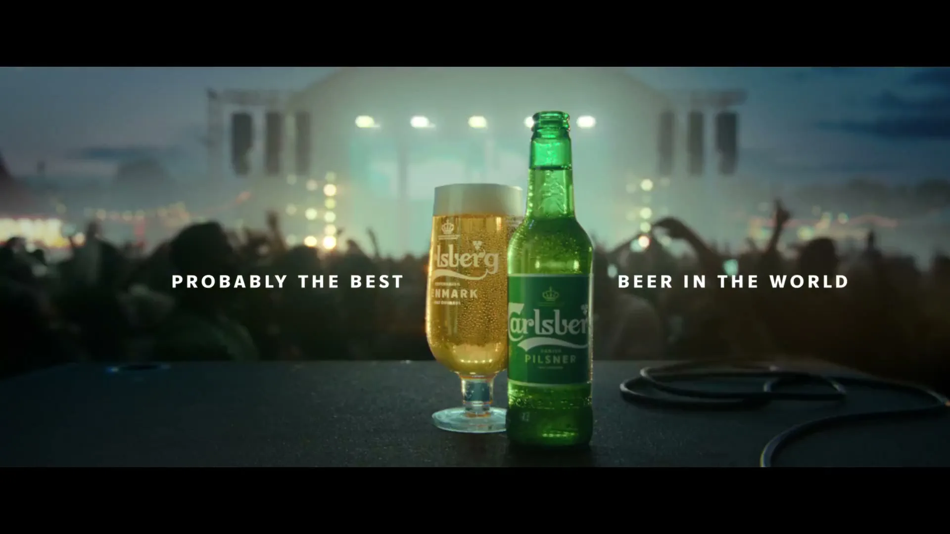 A Carlsberg beer bottle and a glass of beer sit on a dark surface, with a blurred crowd and stage lights in the background at a concert.