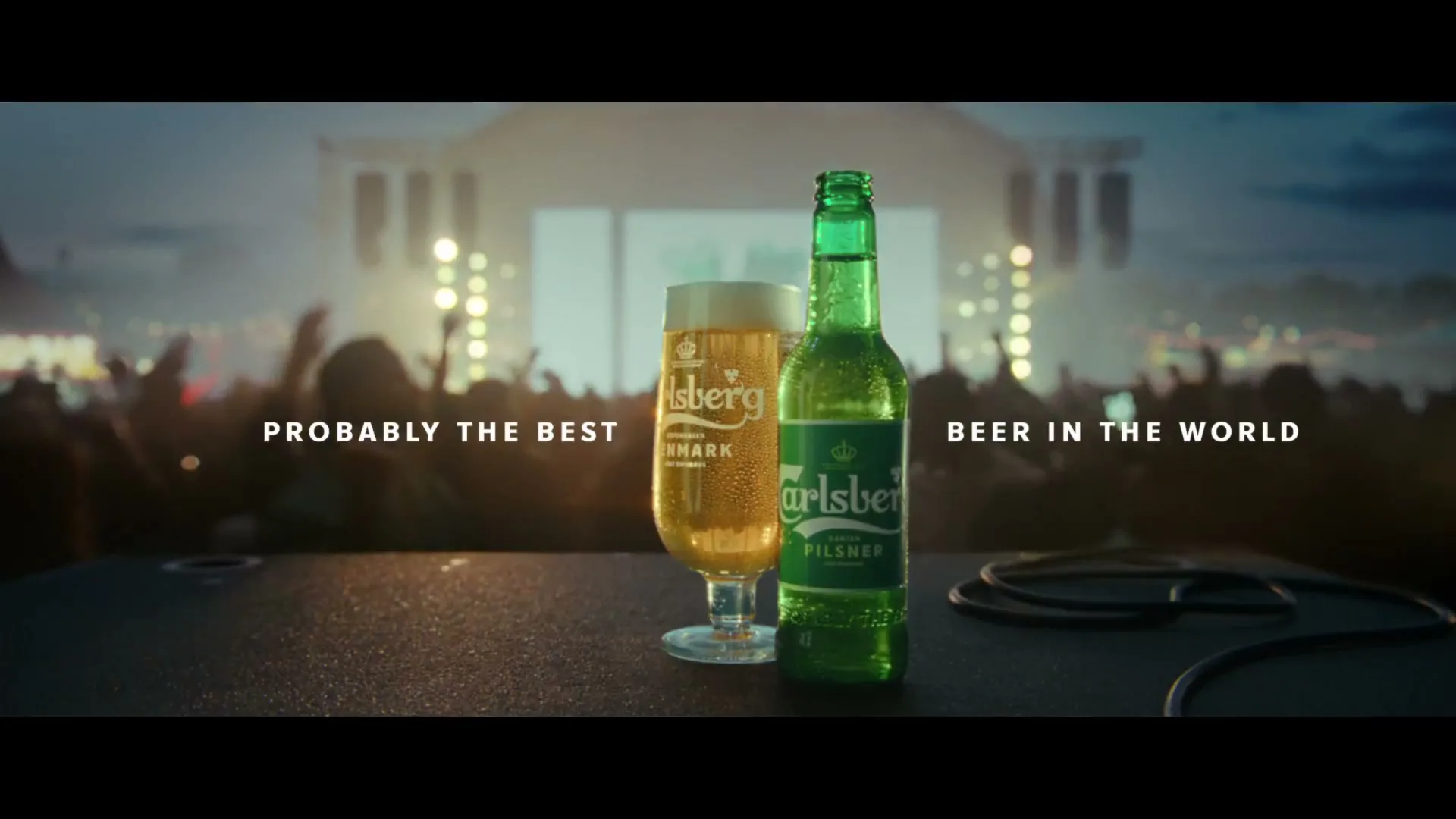 A Carlsberg beer bottle and a glass of beer sit on a stage, with a blurred crowd and concert lights in the background.