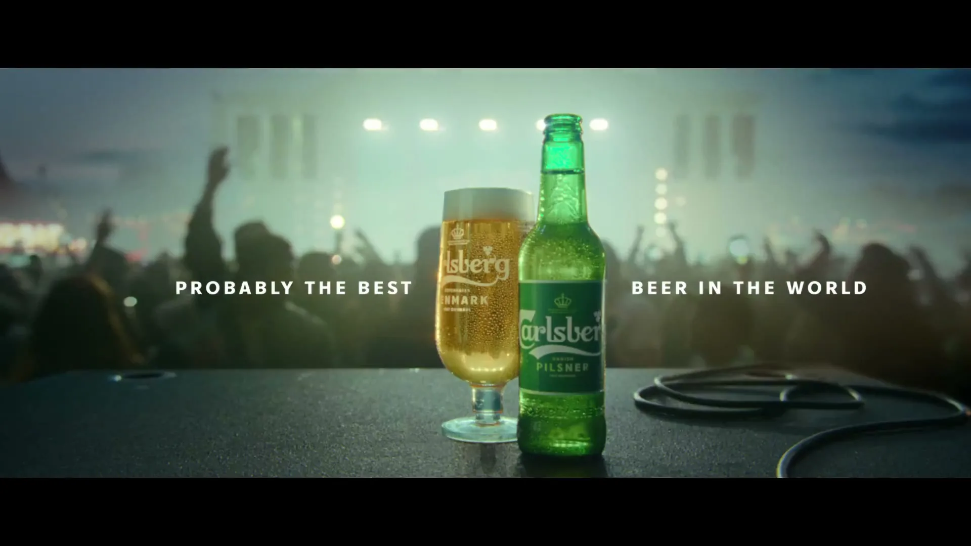 A Carlsberg beer bottle and glass sit on a table in front of a cheering crowd at an outdoor concert at night.