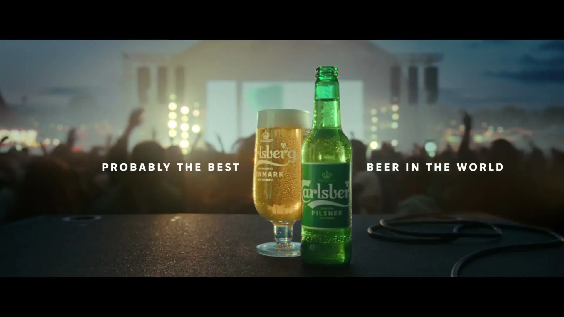 A Carlsberg beer bottle and a glass of beer are on a stage, with a blurred crowd and concert lights in the background.