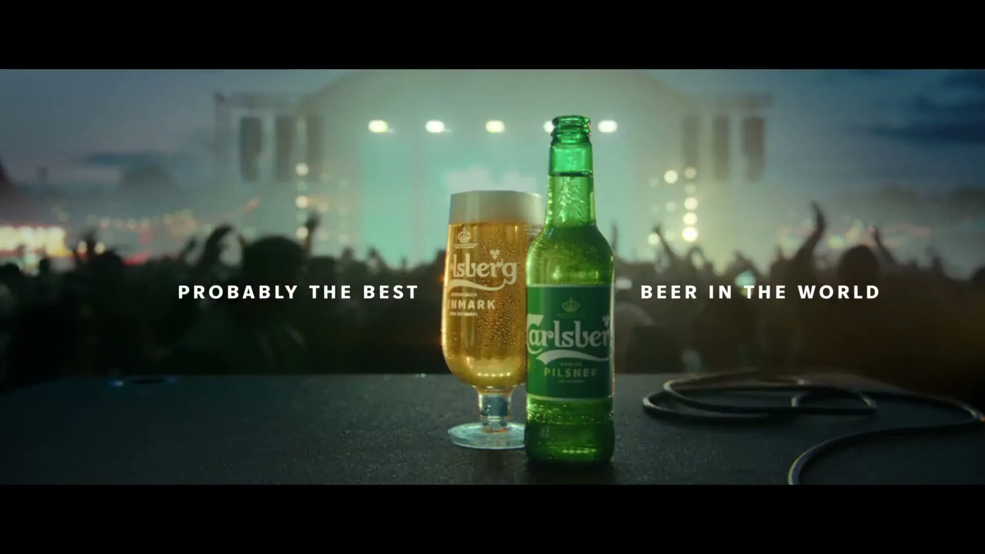 A Carlsberg beer bottle and a glass of beer sit on a stage with a blurred crowd and concert lights in the background.