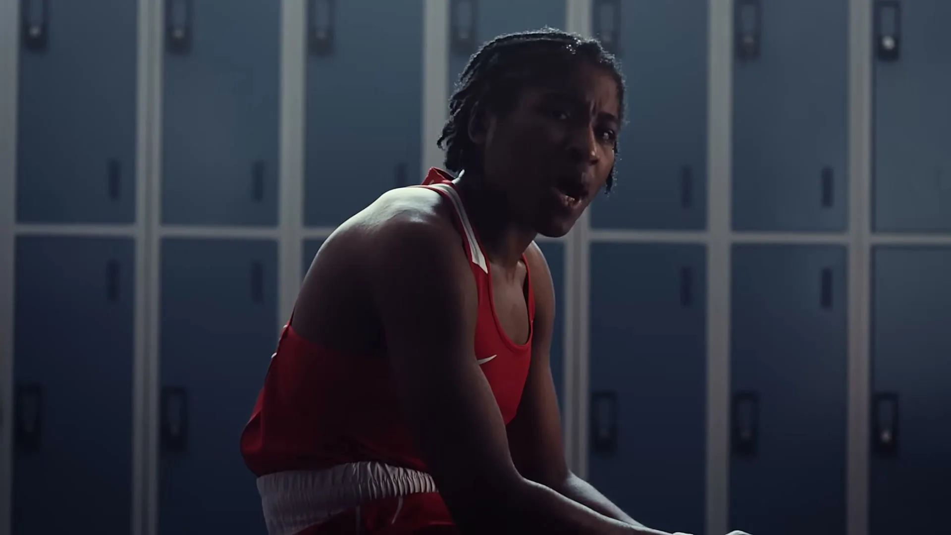 A young female boxer in a red uniform sits on a bench in a locker room, looking intently at the camera.