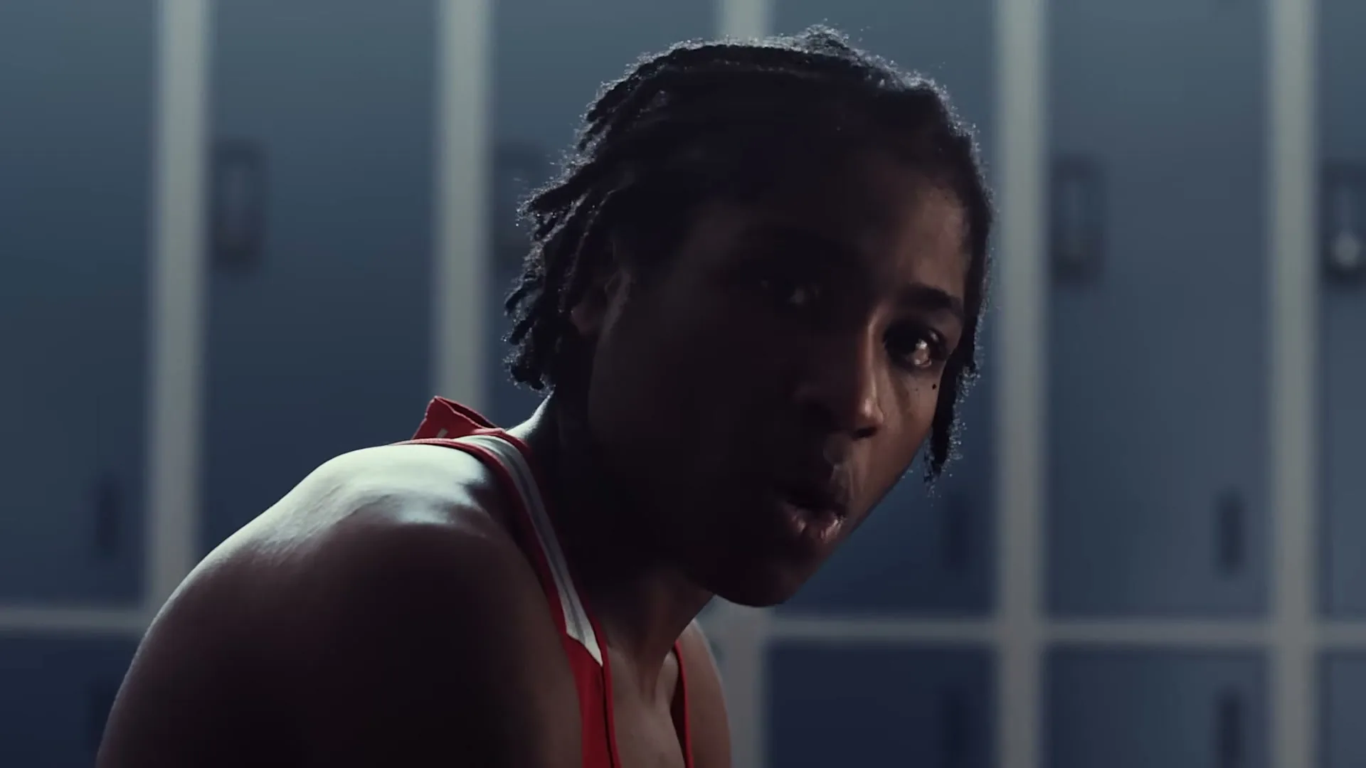 A young Black woman with braided hair, wearing a red sports top, looks intently at the camera in a dimly lit locker room.