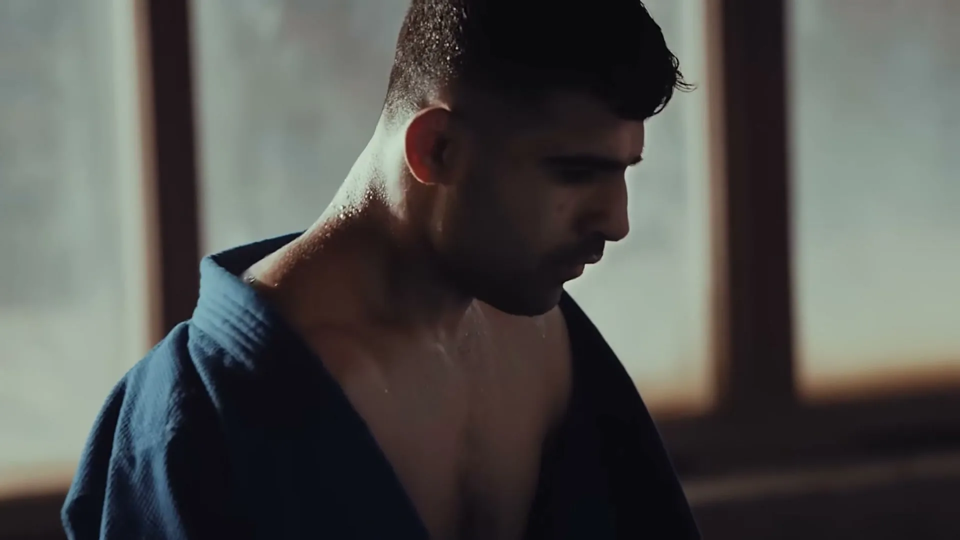 A sweaty male athlete in a blue gi looks down, catching his breath in a sunlit gym.