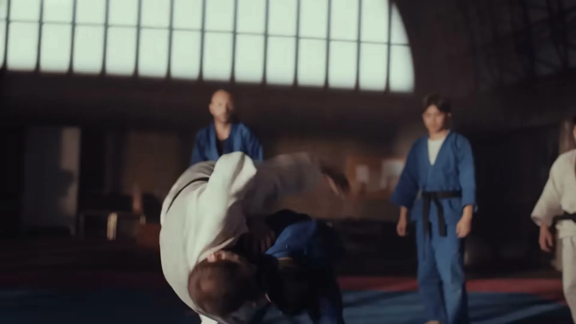 Two male judo athletes in blue and white gis grapple and one throws the other to the mat in a dimly lit gym.
