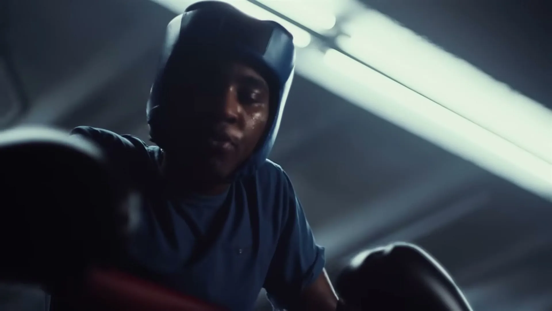 A young male boxer, wearing a blue headguard and boxing gloves, looks up from inside a boxing ring in a dimly lit gym.