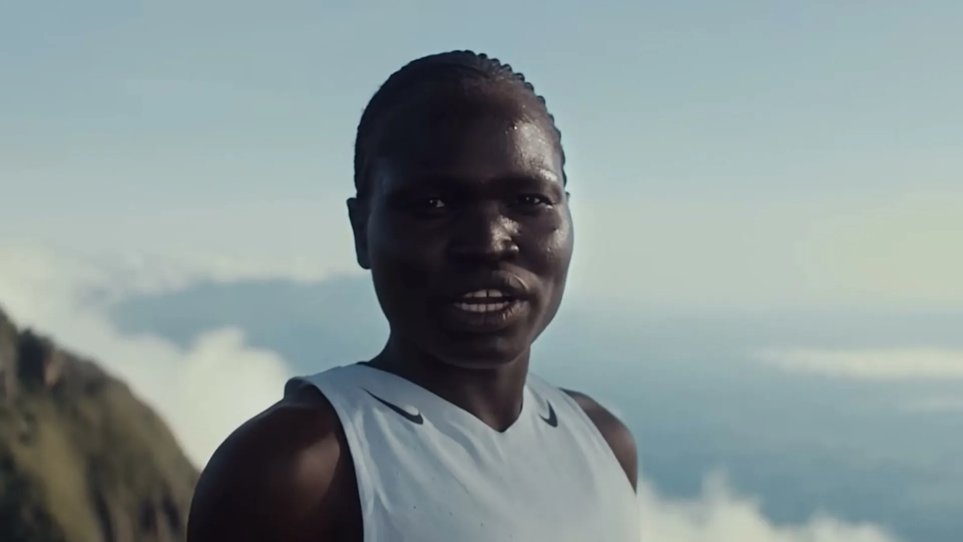 A dark-skinned female athlete with braided hair, wearing a white tank top, looks directly at the camera and speaks on a mountain top with clouds below.