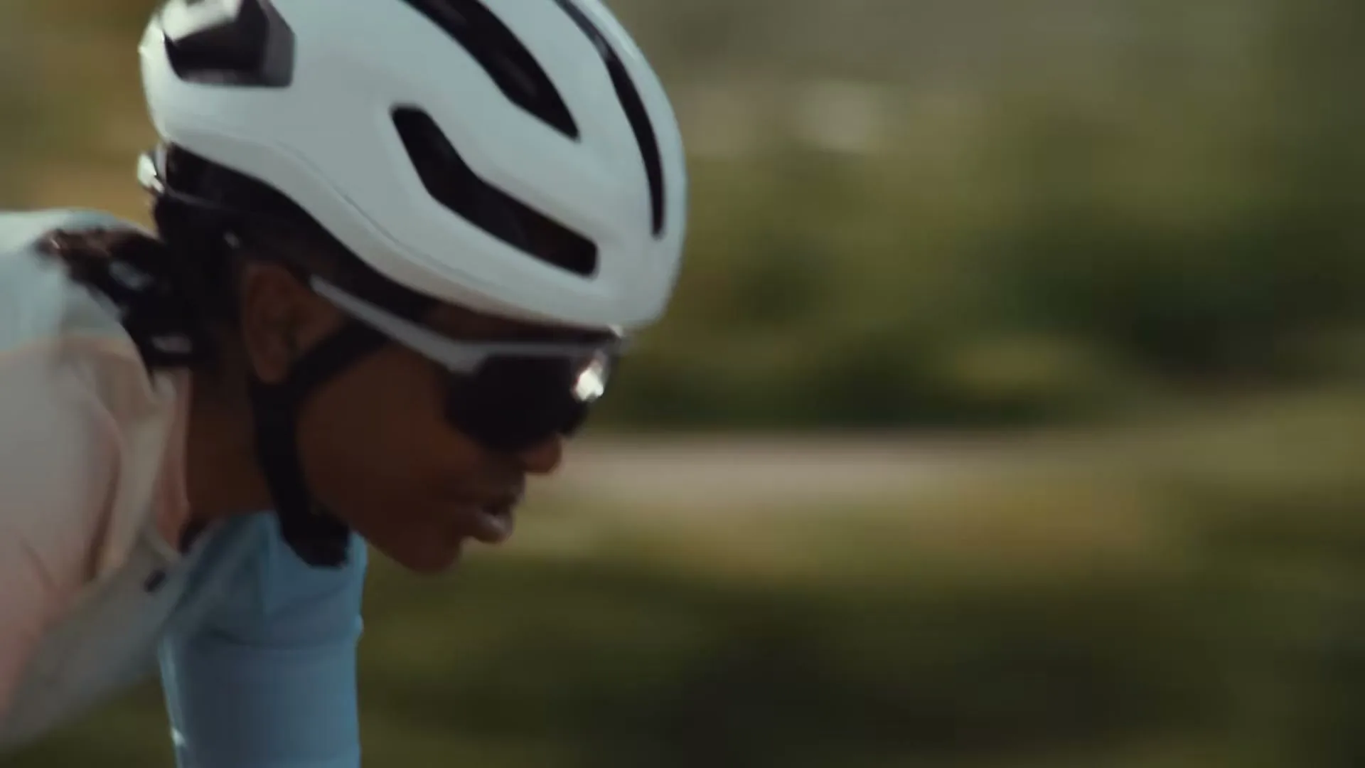 A female cyclist in a white helmet and sunglasses rides a road bike on a paved road, with a blurred green background.