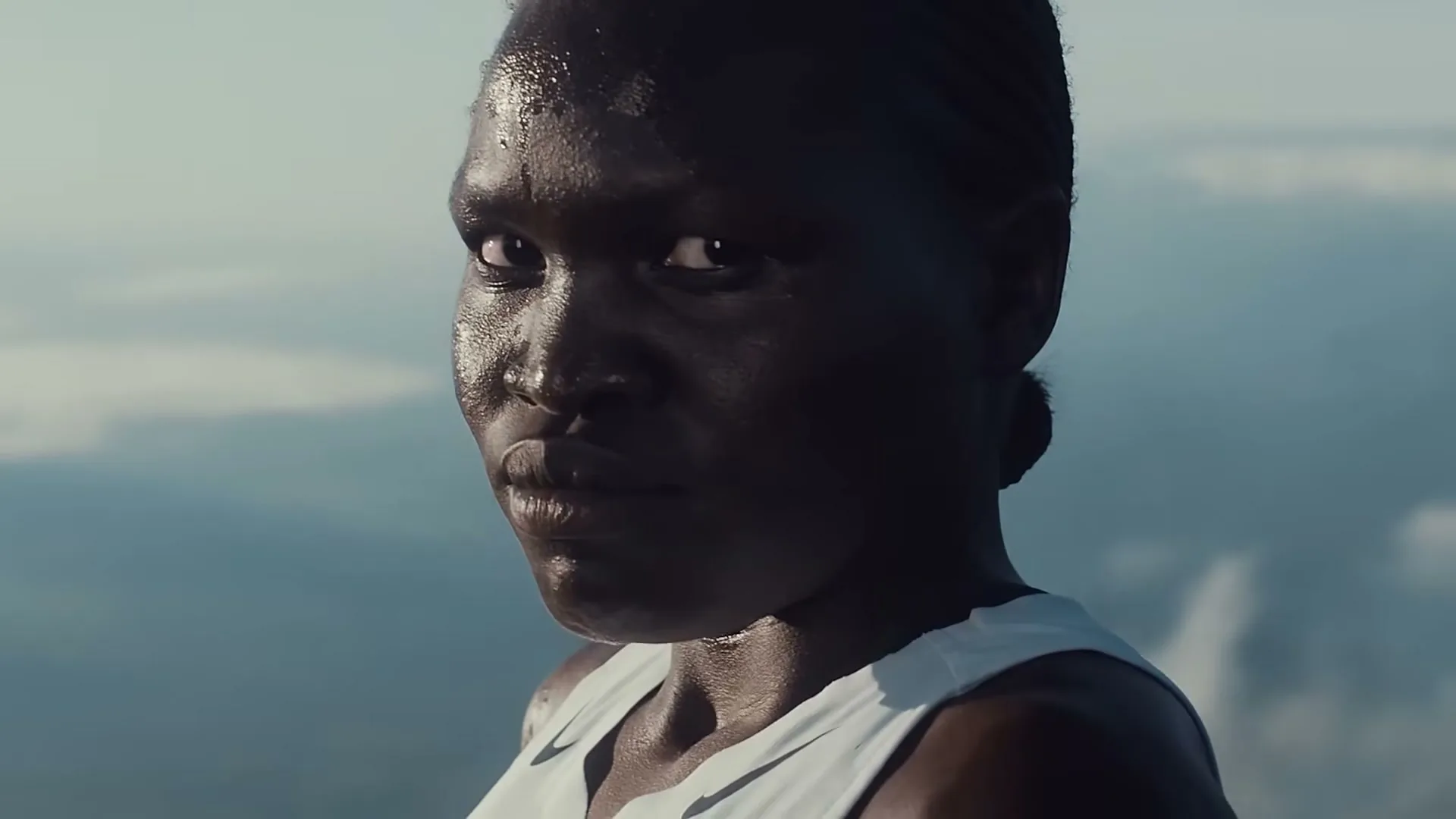A dark-skinned woman with a determined expression, wearing a white tank top, looks directly at the camera with sweat on her face, against a blurry sky background.