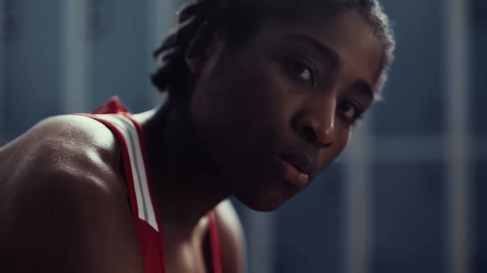 A young Black woman with braided hair, wearing a red sports bra, looks intently at the camera in a locker room.