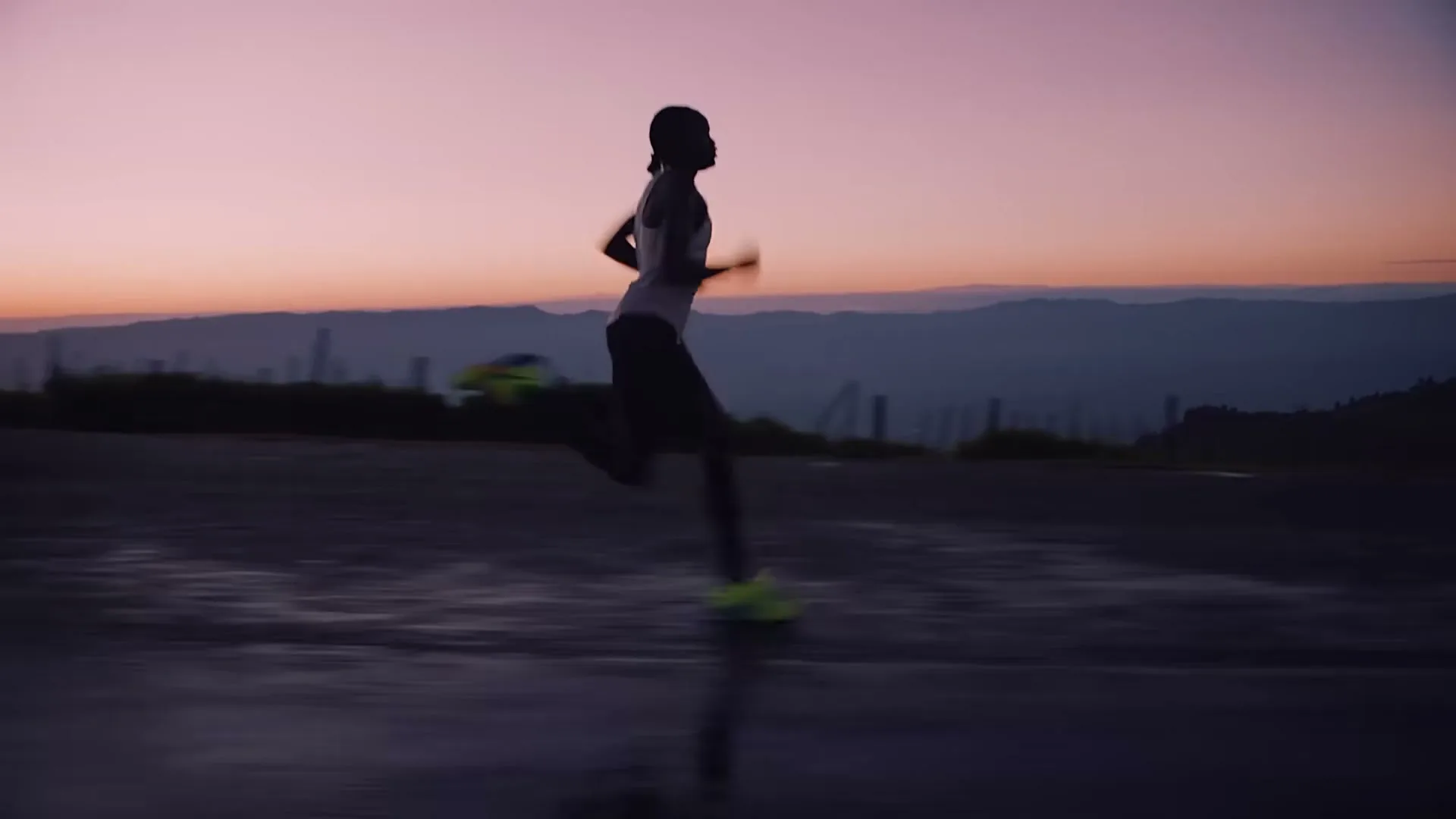 A female runner in a white tank top and dark shorts runs in silhouette against a vibrant pink and purple sunset over mountains.