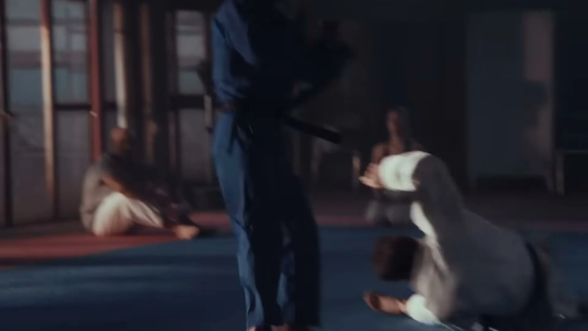 A male athlete in a blue gi throws another male athlete in a white gi onto a blue mat in a dimly lit gym.