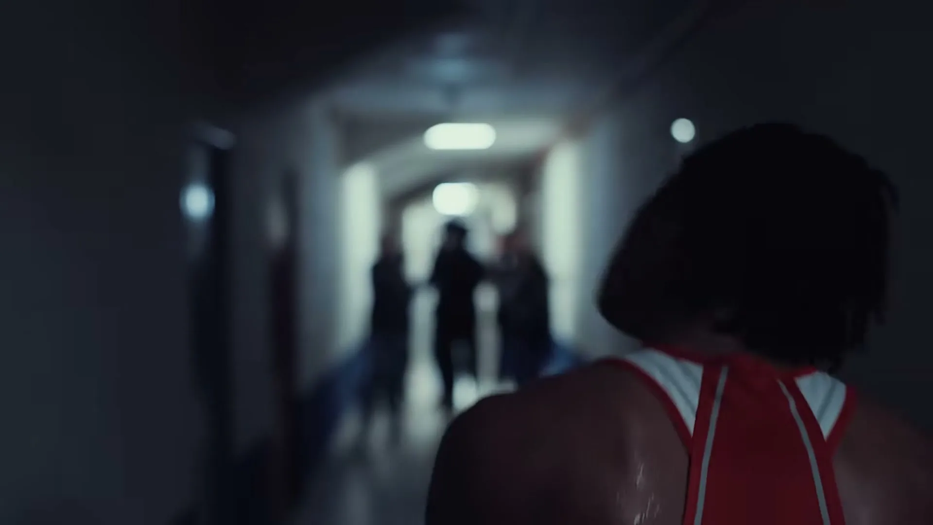 A male athlete with dreadlocks, wearing a red and white singlet, walks away from the camera down a dark, blurry hallway towards a group of people.