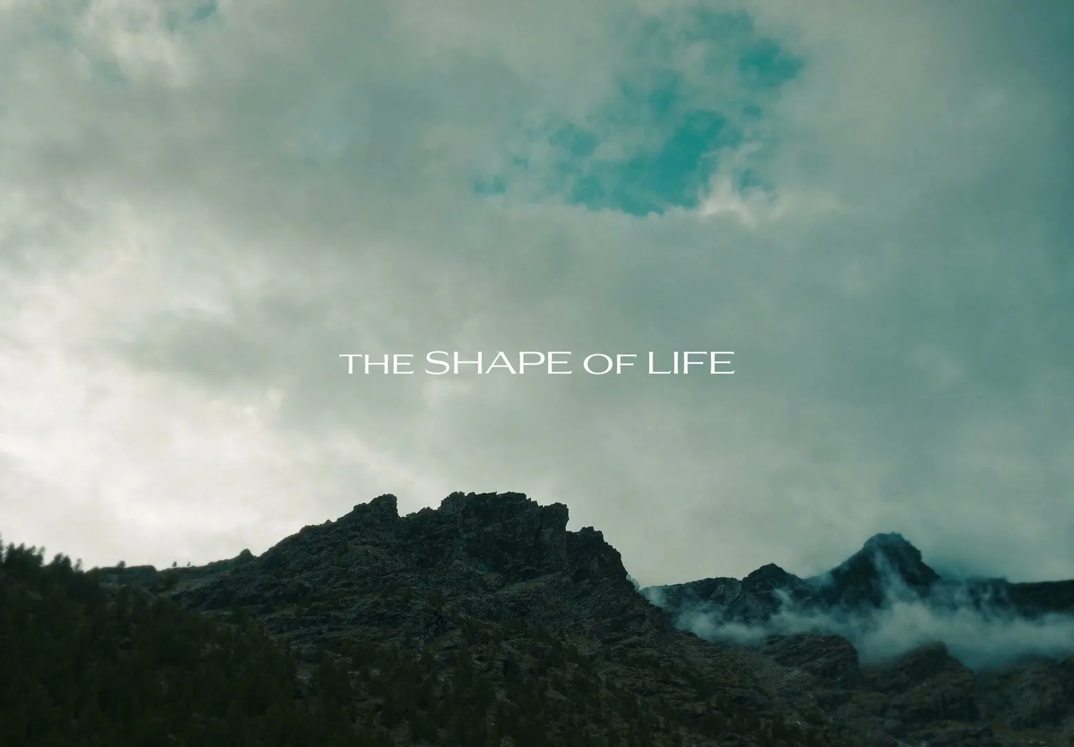 A wide shot of a mountain range under a cloudy sky, with the text "THE SHAPE OF LIFE" superimposed in the center.