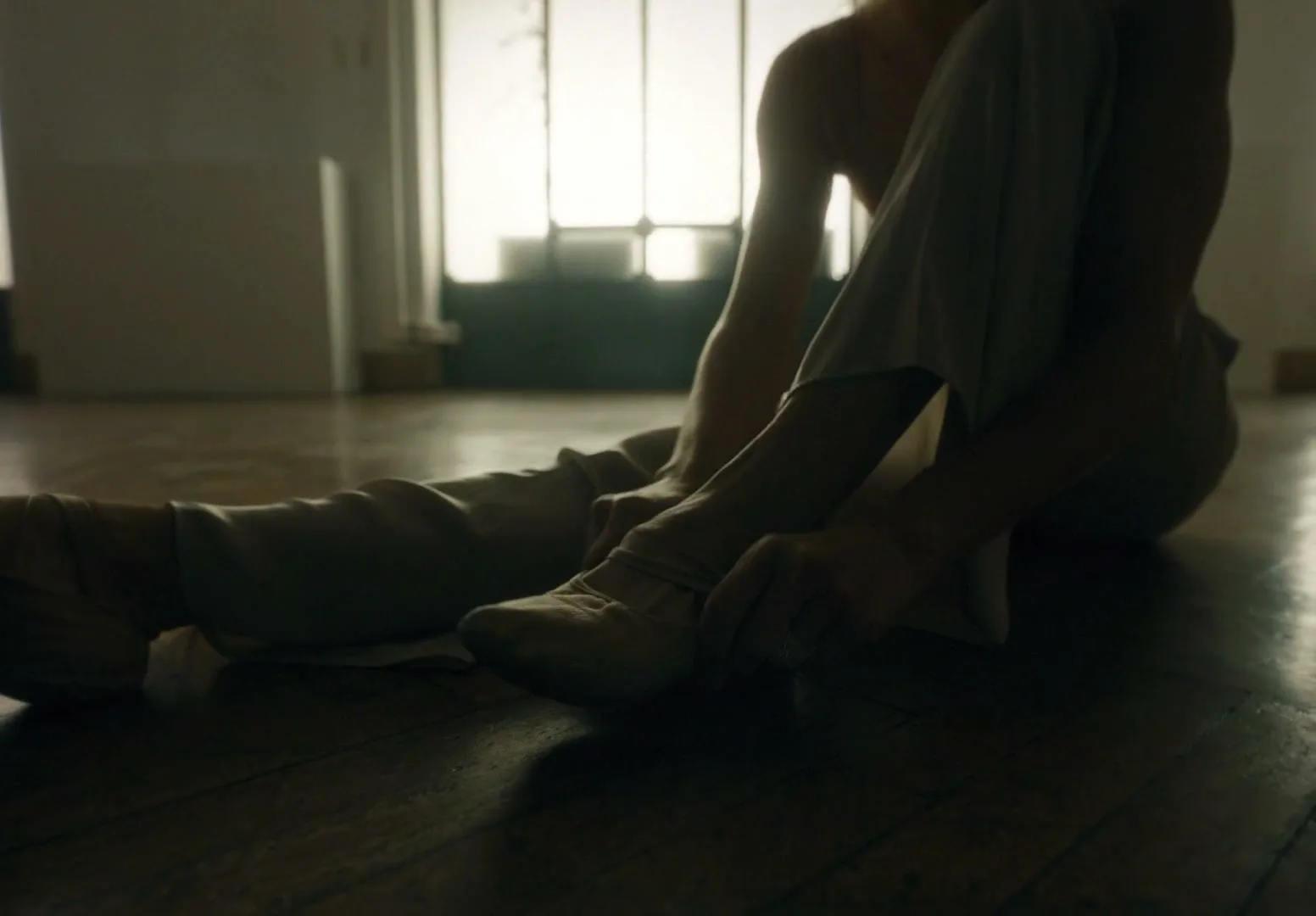 A female dancer sits on a wooden floor, adjusting her ballet slipper, with a bright window in the background of a dance studio.