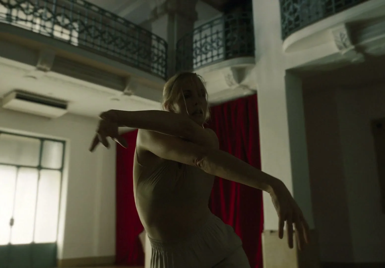 A female dancer in light clothing performs a dramatic movement in a dimly lit, ornate room with red curtains and balconies.