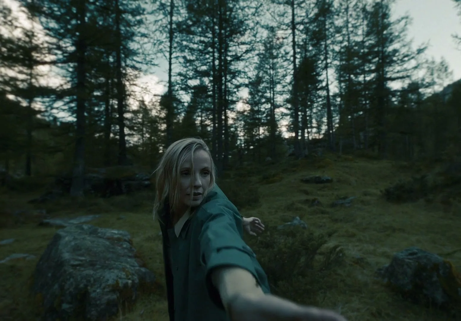 A blonde woman in a green jacket reaches out to the camera, looking back over her shoulder in a dark, mossy forest.