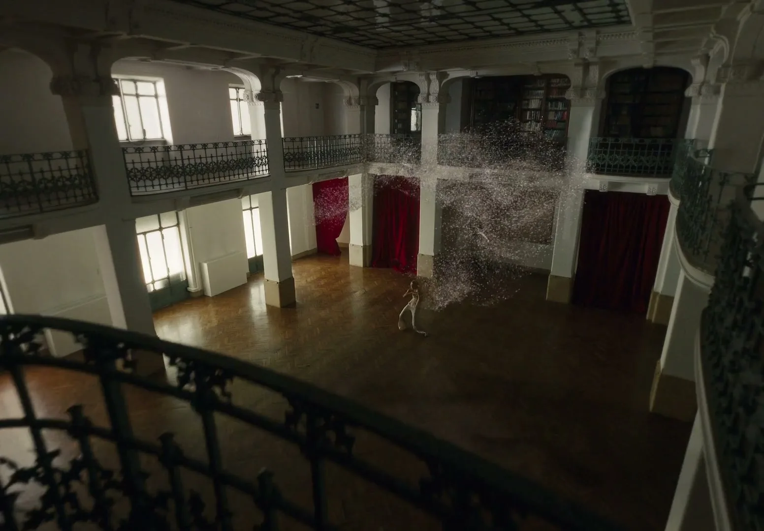 A woman in a white dress dances in a grand, empty hall with balconies and red curtains, surrounded by a swirling cloud of sparkling particles.