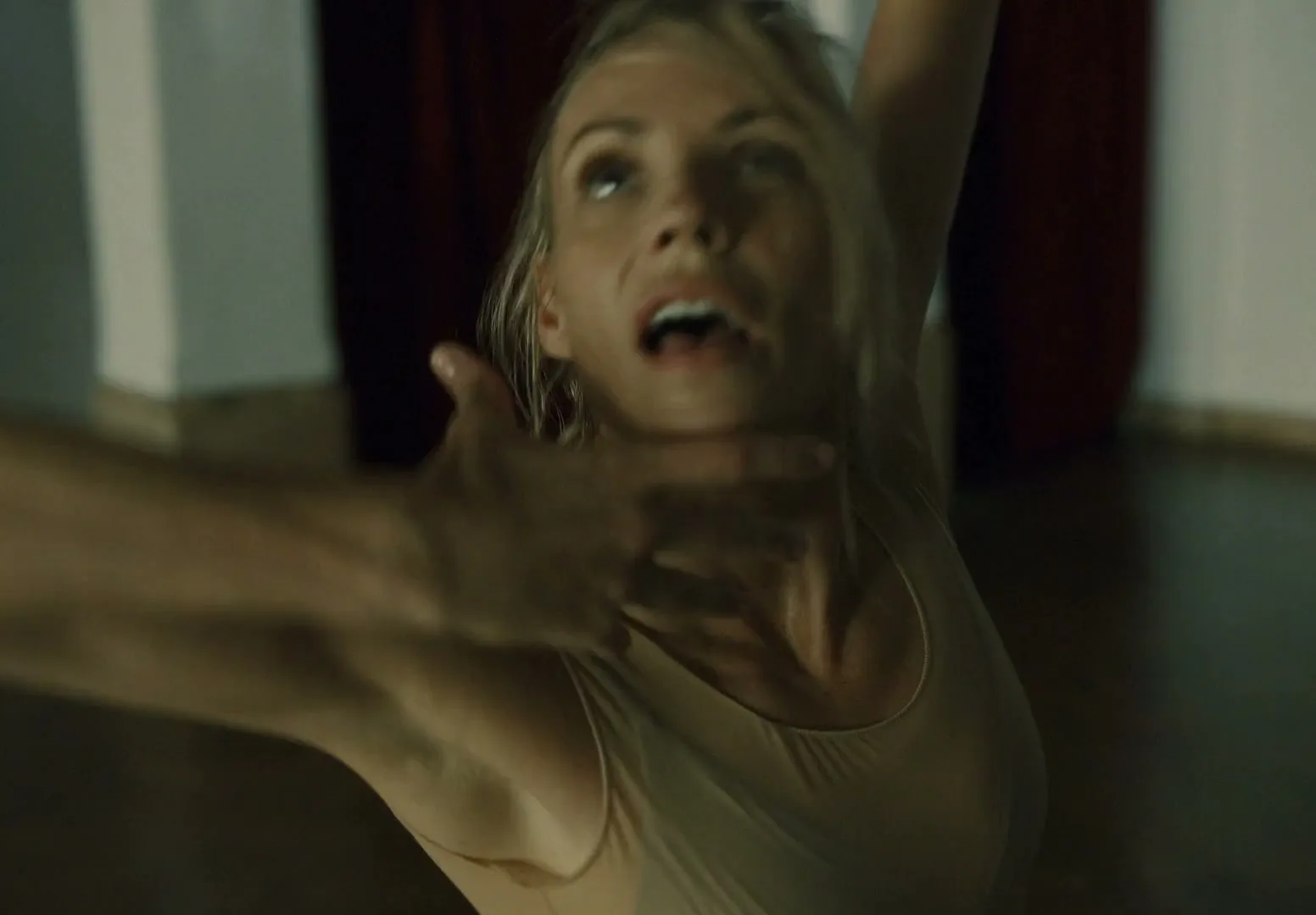 A blonde woman in her 30s, wearing a nude leotard, looks up with a surprised expression as a man's hands move around her face in a dimly lit studio.
