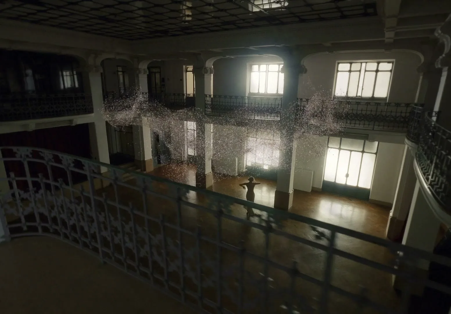 A woman stands in a grand, empty hall with high ceilings and large windows, surrounded by a swirling cloud of sparkling particles.