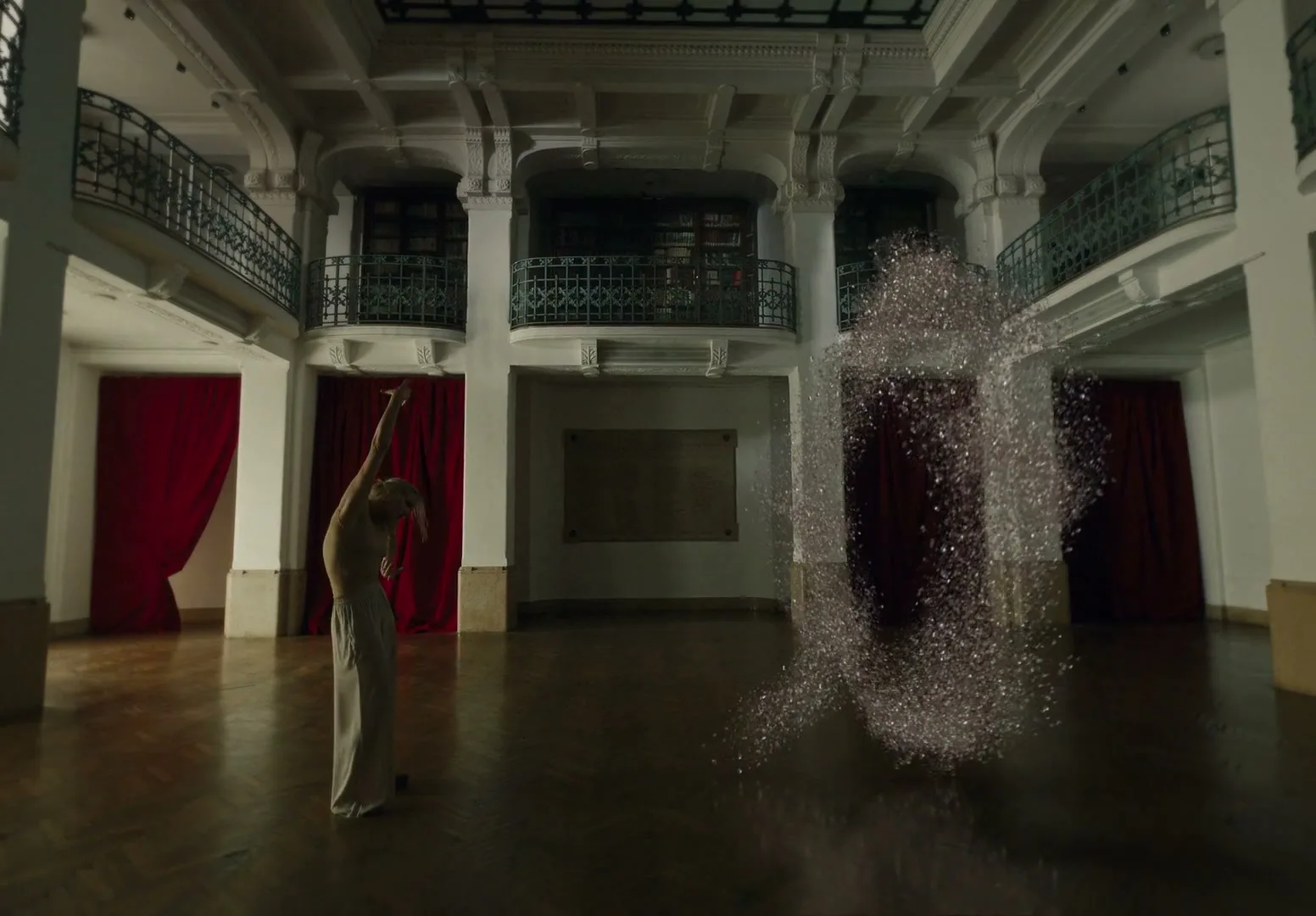 A woman in light clothing dances gracefully in a grand hall, as a shimmering, abstract form of light particles appears beside her.