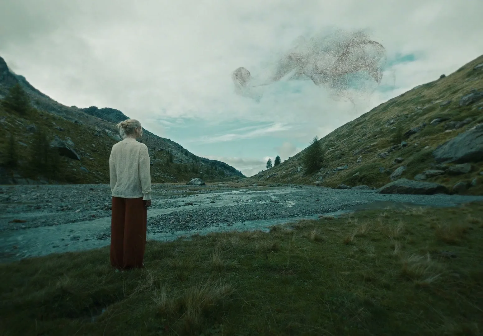 A woman stands in a valley, looking at a swirling mass of particles in the sky above a river.