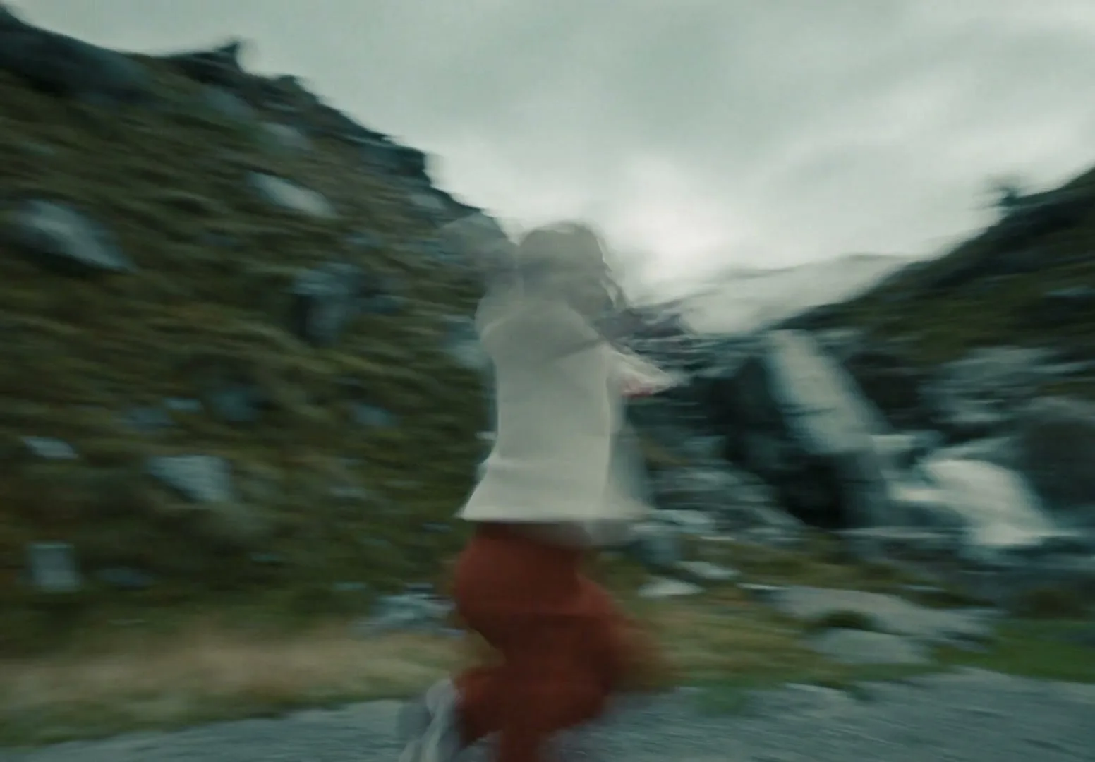 A blurry shot of a woman in a white sweater and red pants running through a rocky, grassy mountain pass under a cloudy sky.