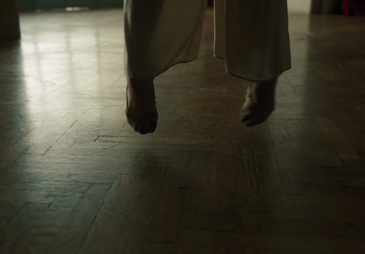 A person's feet in light-colored pants and shoes are seen from the knees down, jumping on a wooden floor in a dimly lit room.