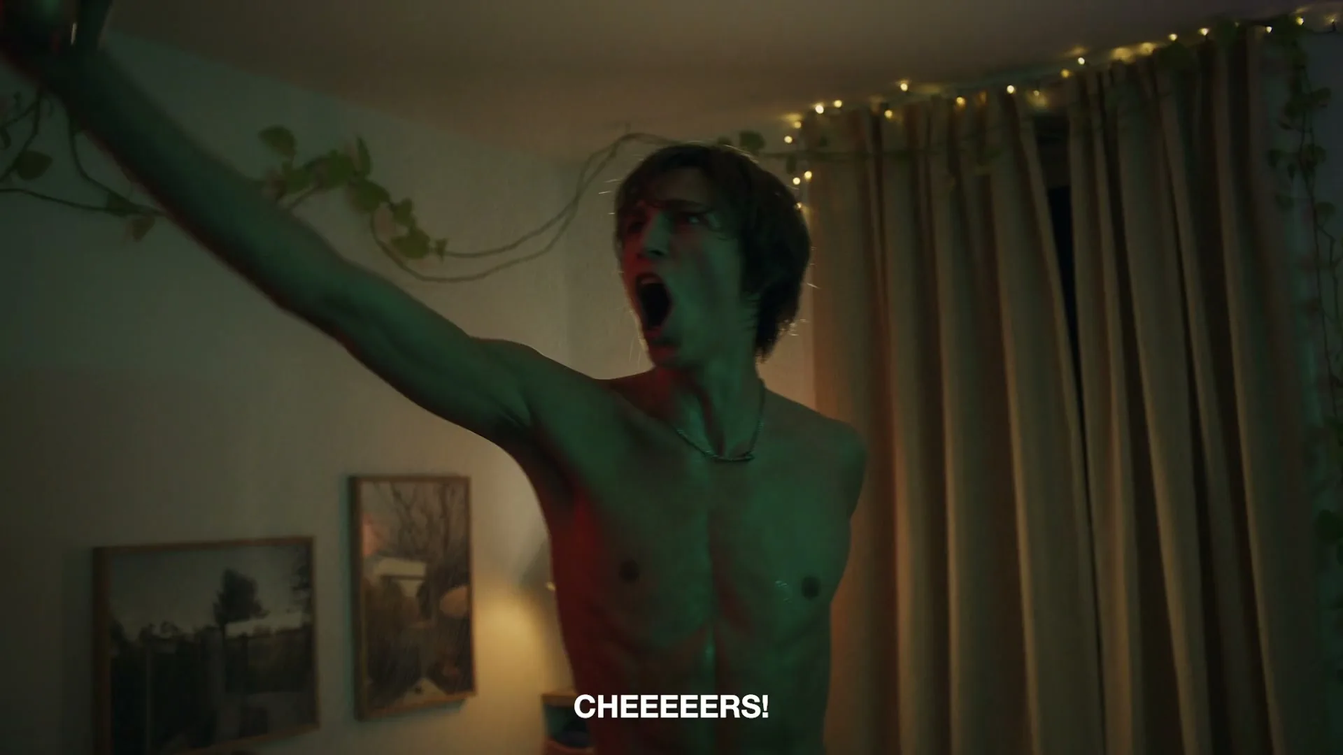 A shirtless young man with long hair sprays beer from a can into his mouth, then shouts with his arm raised at a dimly lit house party.