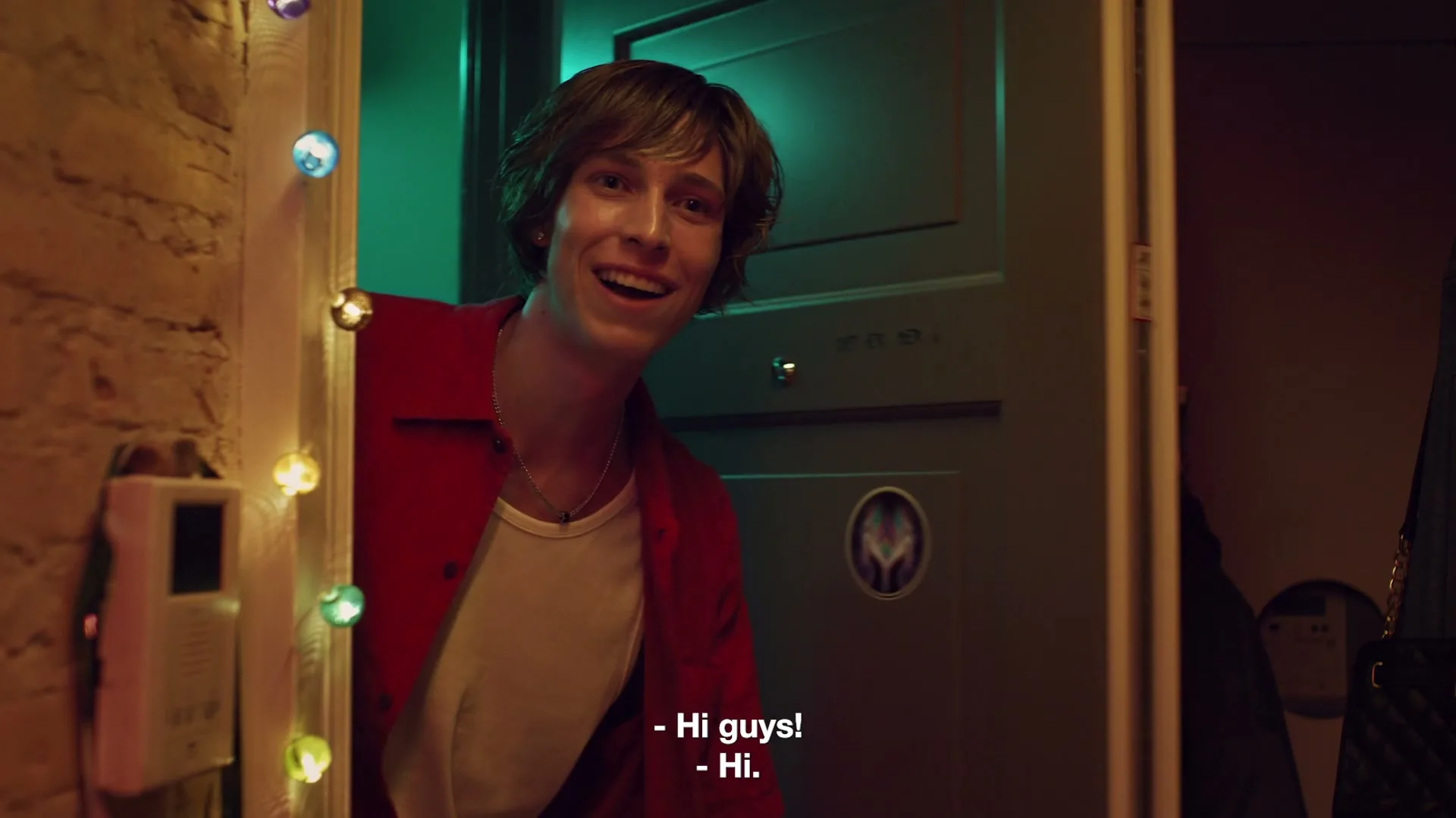 A young man with brown hair, wearing a red shirt and white undershirt, peeks out from behind a door, smiling and saying "Hi guys!" in a dimly lit hallway decorated with string lights.